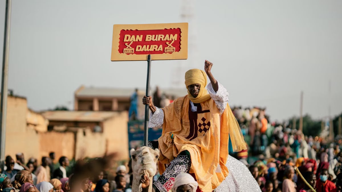 A vibrant cultural parade scene with a traditional figure raising a sign in Daura, Nigeria.